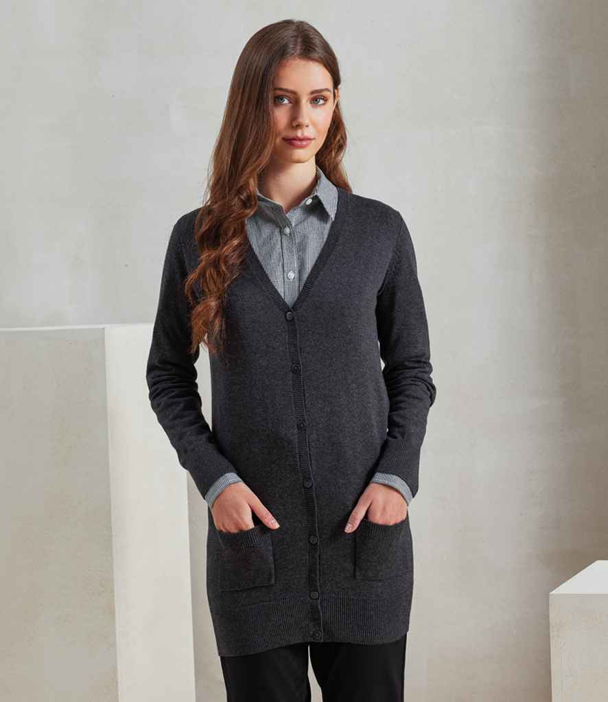 A woman with long brown hair stands indoors, hands in her pockets, wearing the Premier Ladies Longline V Neck Cardigan by Premier over a gray collared shirt and black pants against a plain, neutral background.