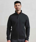 A man with short dark hair and a beard stands against a light background, wearing the Premier Spun Dyed Recycled Zip Through Sweat Jacket by Premier with a high collar and dark pants. He faces forward with a neutral expression.
