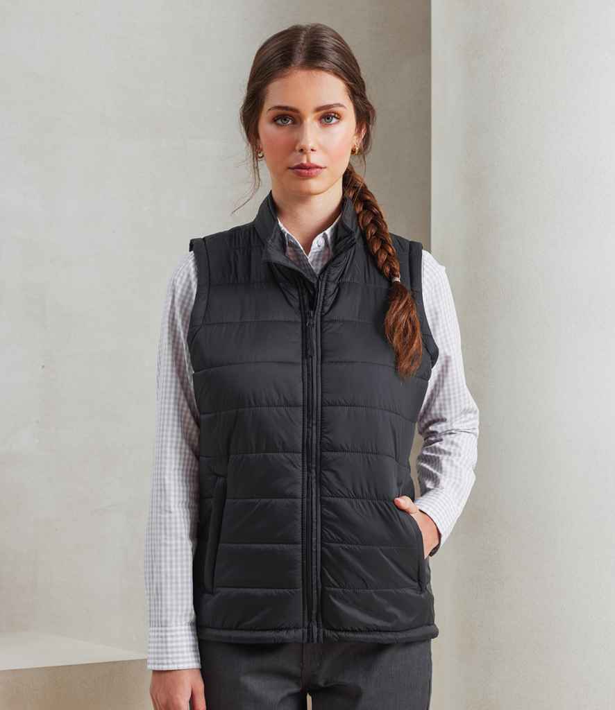 A woman stands indoors facing the camera against a neutral background, wearing the Premier Ladies Recyclight® Padded Gilet by Premier—a black, shower-resistant, recycled polyester vest—over a light checked shirt and dark trousers, with a long braid over her shoulder.