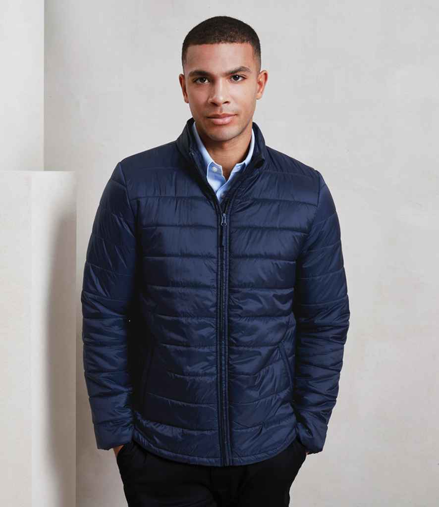 A person stands against a plain background wearing the Premier Recyclight® Padded Jacket by Premier—dark blue, lightweight, made from recycled polyester—over a light blue collared shirt, hands relaxed at their sides with a neutral expression.