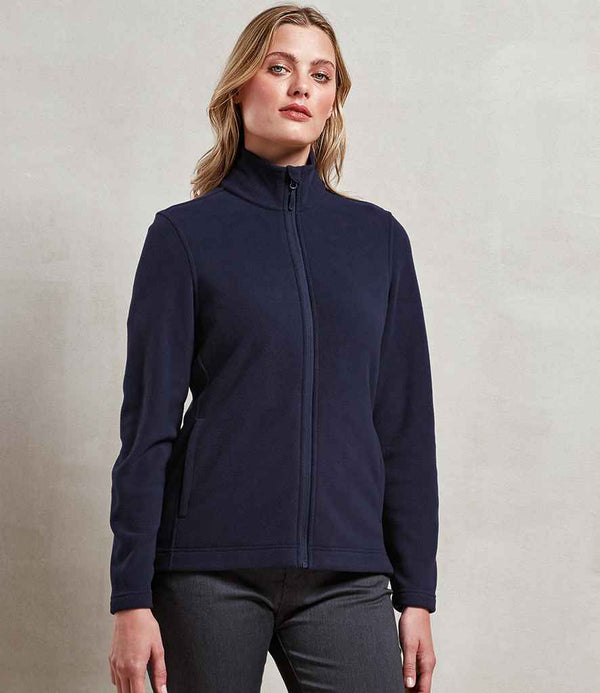 A person stands against a plain light wall, wearing the Premier Ladies Recyclight® Full Zip Micro Fleece Jacket by Premier in navy with a cadet collar, full zip, and anti-pill finish. They pair it with dark pants and face forward neutrally.