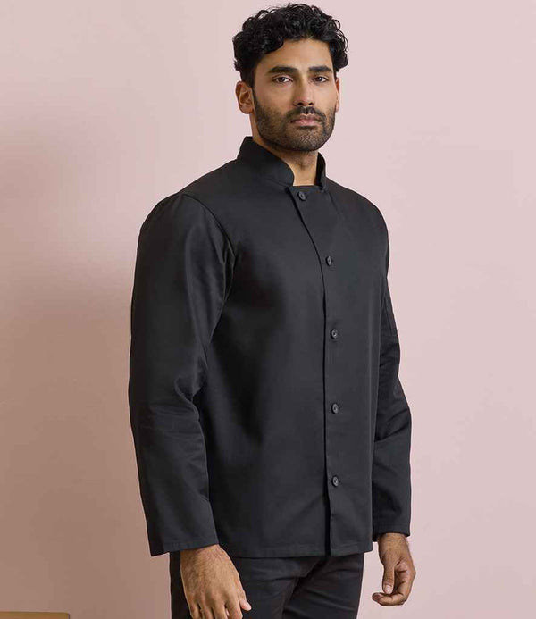 A man with short, curly hair and a trimmed beard stands against a pale pink background wearing the Premier Premier Essential Long Sleeve Chefs Jacket in black, featuring a Mandarin collar and resin buttons down the front.