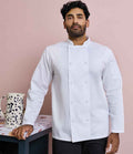 A dark-haired, bearded man wears a Premier Coolchecker® Long Sleeve Chefs Jacket by Premier with a mandarin collar. He stands next to a table holding a large ceramic jug, set against a light pink background.