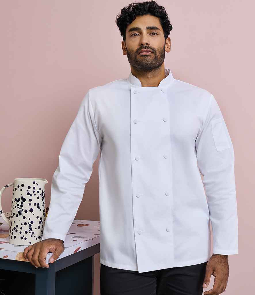 A dark-haired, bearded man wears a Premier Coolchecker® Long Sleeve Chefs Jacket by Premier with a mandarin collar. He stands next to a table holding a large ceramic jug, set against a light pink background.