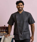 A man with short curly hair and a beard smiles while standing beside a counter with baked goods, wearing the Premier Short Sleeve Zipped Chefs Jacket from Premier. The background is light pink.
