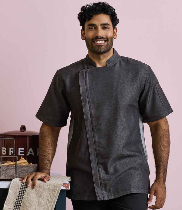 A man with short curly hair and a beard smiles while standing beside a counter with baked goods, wearing the Premier Short Sleeve Zipped Chefs Jacket from Premier. The background is light pink.