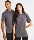 Two people wear matching Premier Essential Unisex Polo Shirts by Premier, featuring short sleeves and a ribbed collar, standing side by side against a plain background. One smiles and looks away; the other faces forward with a neutral expression.