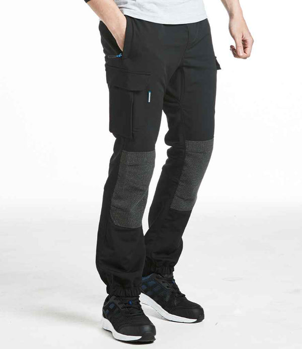 Someone wearing Portwest KX3™ Flexi Trousers by Portwest, featuring a slim fit and reinforced knees, paired with black sneakers, stands hands-in-pockets against a plain white background.