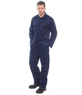 A man stands facing forward against a plain white background, wearing the Portwest Euro Work Coverall in dark blue with chest pockets and an elasticated waistband from Portwest, paired with black work boots and hands in his pockets.