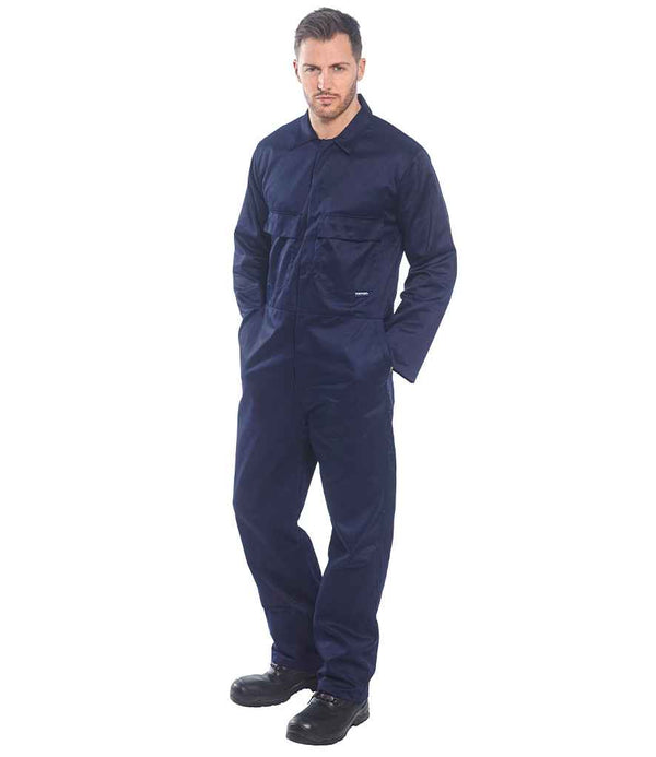 A man stands facing forward against a plain white background, wearing the Portwest Euro Work Coverall in dark blue with chest pockets and an elasticated waistband from Portwest, paired with black work boots and hands in his pockets.