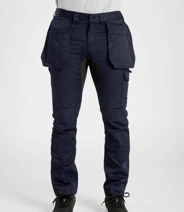 A person wears Portwest WX2™ Eco Stretch Holster Trousers—dark blue with large holster and knee pad pockets—paired with a light shirt and black shoes, shown from the waist down. Brand: Portwest.