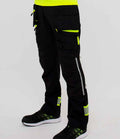 A person wearing Portwest DX4™ Detachable Holster Pocket Trousers by Portwest, featuring multiple pockets and knee pad pockets, along with black and green athletic shoes, stands against a plain light background.