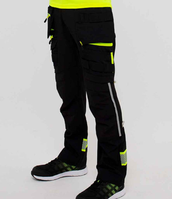 A person wearing Portwest DX4™ Detachable Holster Pocket Trousers by Portwest, featuring multiple pockets and knee pad pockets, along with black and green athletic shoes, stands against a plain light background.