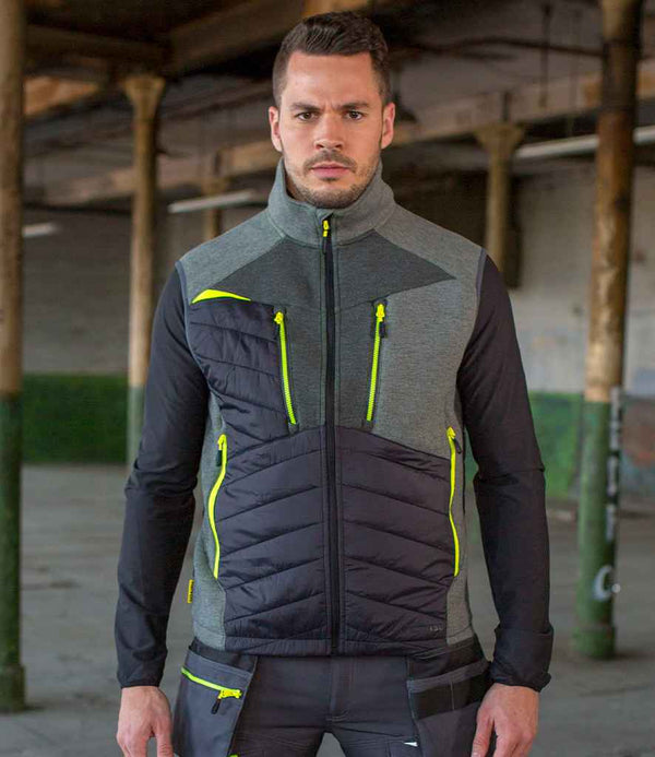 A man with short dark hair and a trimmed beard stands indoors wearing the Portwest DX4™ Baffle Gilet, a gray and black zip-up vest with neon yellow accents and reflective piping, in an industrial setting with pillars and an exposed ceiling.