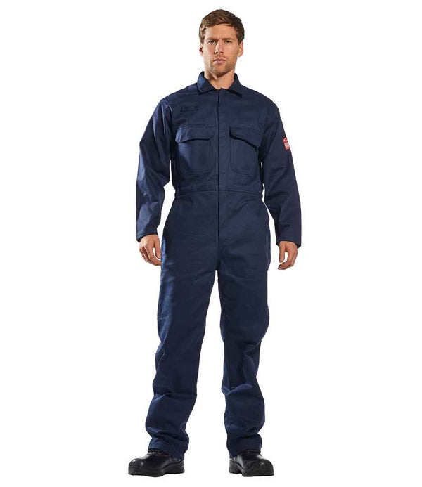 A person faces forward in a Portwest Bizweld™ Flame Resistant Coverall by Portwest—navy blue, long sleeves, chest pockets—and black work boots. The background is plain white.