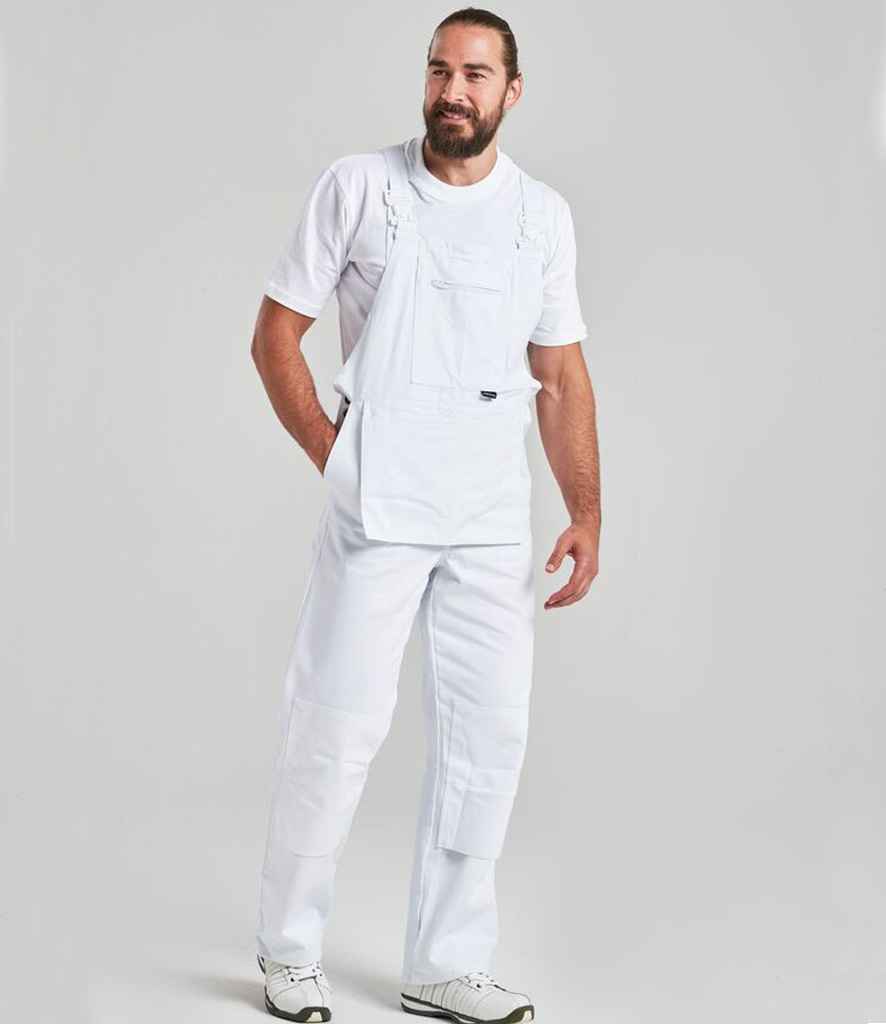 A man with a beard wears the Portwest Bolton Painters Bib N Brace by Portwest over a white t-shirt and white sneakers, standing against a plain gray background with one hand in his pocket.