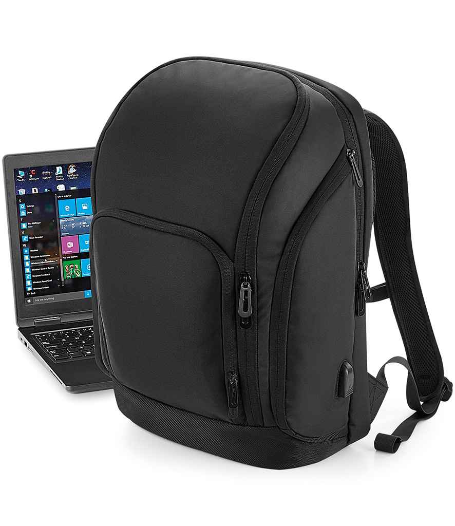 The Quadra Pro-Tech Charge Backpack by Quadra, featuring multiple zippered compartments, an ergonomic back panel, padded straps, and a side USB charging port, stands upright next to an open laptop displaying Windows.