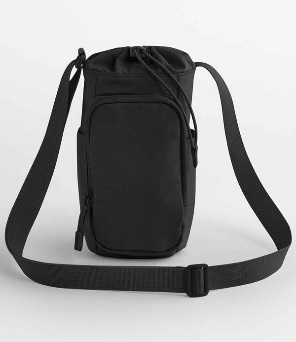 The Quadra Studio Water Bottle Carrier by Quadra is a compact black crossbody bag with an adjustable strap, front zip pocket, and top drawstring closure made from water repellent fabric, shown against a plain white background.