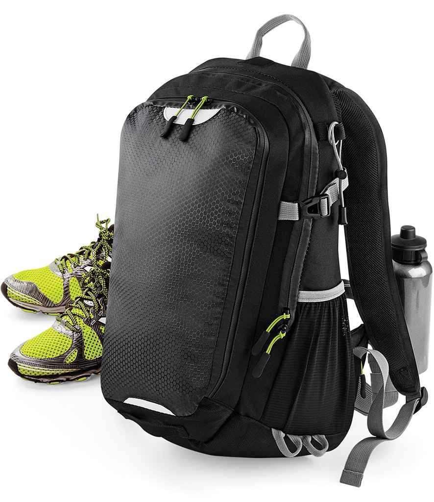 The Quadra SLX 20 Litre Daypack by Quadra, a black and gray waterproof backpack with green zipper pulls, stands upright. Yellow and gray running shoes sit beside it; a silver water bottle rests in the side pocket. Its hydration bladder compatible.