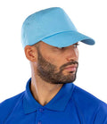 A bearded man wears a light blue Result Cotton Cap by Result Headwear, featuring an adjustable fit, and a blue collared shirt. He looks to his left against a plain white background.