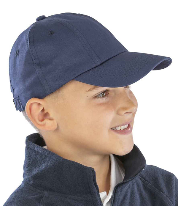 A young boy is shown in profile wearing the Result Kids Low Profile Cotton Cap by Result Headwear and a navy zip-up jacket. He faces right, smiling, against a plain white background.