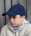 A boy wears the Result Kids Low Profile Heavy Brushed Cotton Cap by Result Headwear, a light gray hoodie, and a beige knit sweater as he sits in front of a wooden fence, looking slightly to the side with a neutral expression.