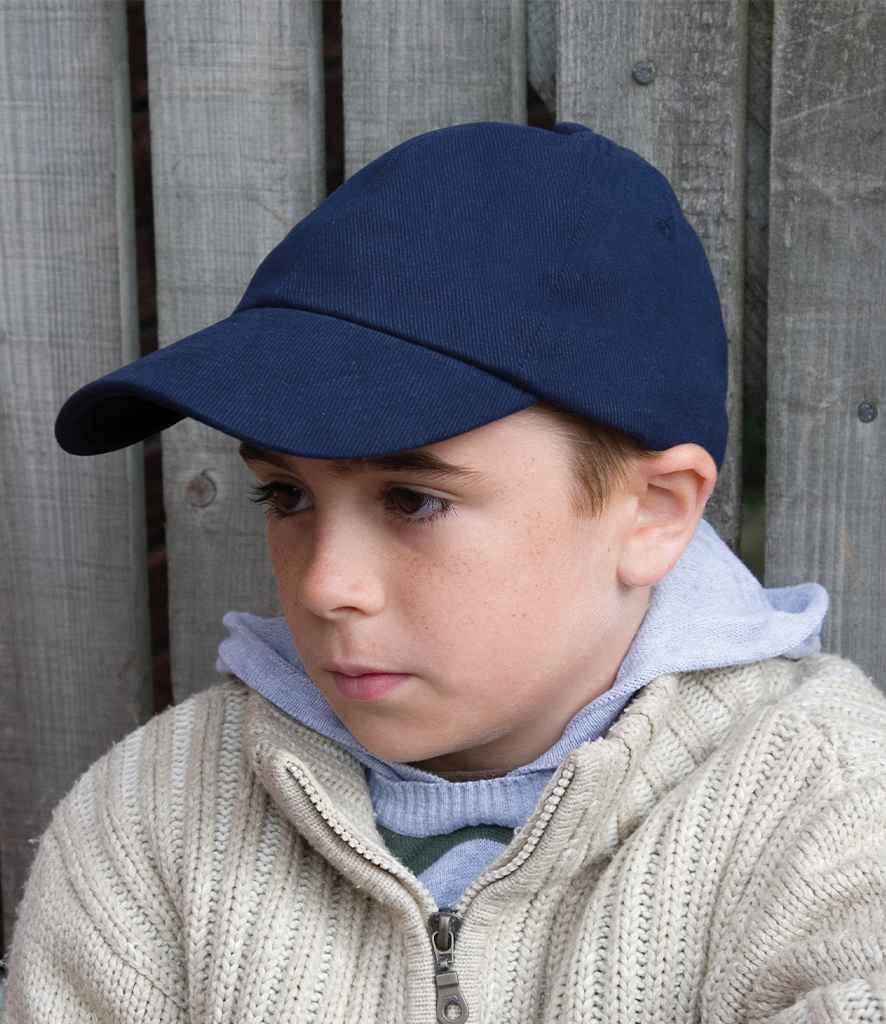 A boy wears the Result Kids Low Profile Heavy Brushed Cotton Cap by Result Headwear, a light gray hoodie, and a beige knit sweater as he sits in front of a wooden fence, looking slightly to the side with a neutral expression.