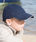 A boy wearing a Result Kids Low Profile Heavy Brushed Cotton Cap with Sandwich Peak by Result Headwear in navy blue and a beige knit sweater sits near water, gazing to the side with a neutral expression.