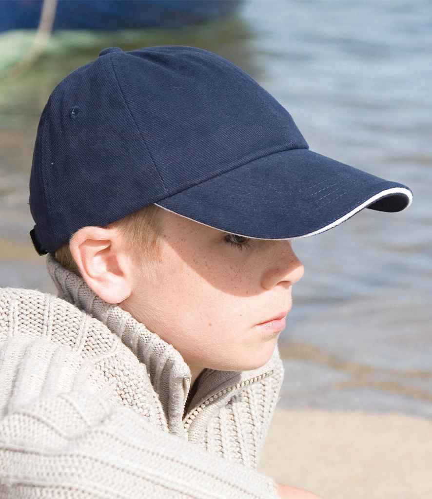 A boy wearing a Result Kids Low Profile Heavy Brushed Cotton Cap with Sandwich Peak by Result Headwear in navy blue and a beige knit sweater sits near water, gazing to the side with a neutral expression.
