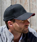 A man with a short beard and mustache wears a Result Headwear Result Pro-Style Heavy Brushed Cotton Cap, featuring six panels, black fabric, and a red-trimmed pre-curved peak. A light gray zip-up jacket and rustic wooden planks complete the scene.
