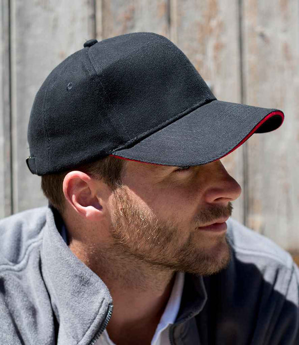 A man with a short beard and mustache wears a Result Headwear Result Pro-Style Heavy Brushed Cotton Cap, featuring six panels, black fabric, and a red-trimmed pre-curved peak. A light gray zip-up jacket and rustic wooden planks complete the scene.