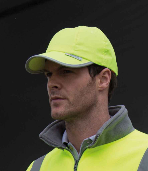 A man with short hair stands against a dark background, wearing a Result Headwear Result High-Viz Cap and a matching safety vest. He looks slightly to the side.