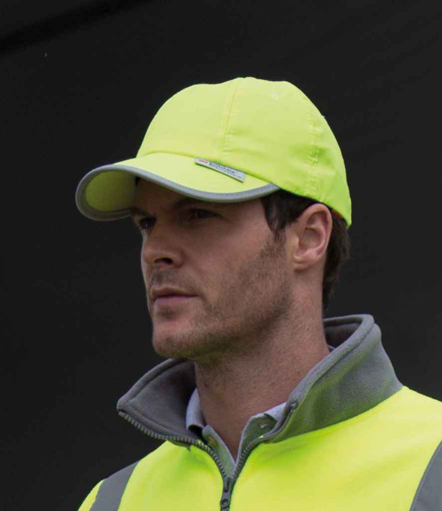 A man with short hair stands against a dark background, wearing a Result Headwear Result High-Viz Cap and a matching safety vest. He looks slightly to the side.