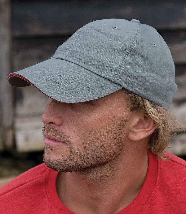 A man with light facial hair and blond hair wears the Result Headwear Result Herringbone Cap in light grey with an adjustable buckle and a red shirt, looking left. The blurred background features wooden planks.