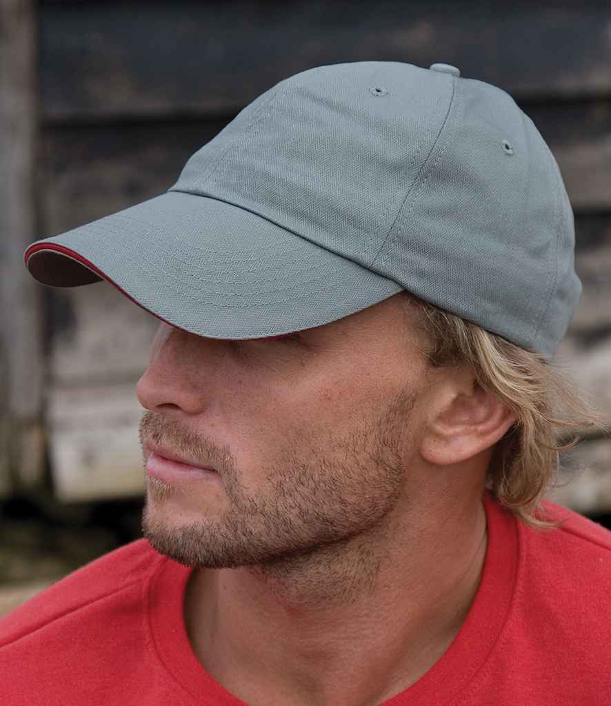 A man with light facial hair and blond hair wears the Result Headwear Result Herringbone Cap in light grey with an adjustable buckle and a red shirt, looking left. The blurred background features wooden planks.