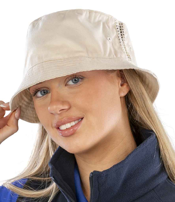 A person with long blonde hair wears the Result Headwear Result Deluxe Washed Cotton Bucket Hat in light beige and a dark zip-up jacket, facing the camera, slightly tilting the hat with one hand. The background is plain white.