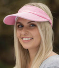 A person with long blonde hair is smiling and looking slightly left while wearing the Result Headwear Result Herringbone Sun Visor, which has a light pink color, pre-curved peak, and gray top. The blurred background is outdoors.