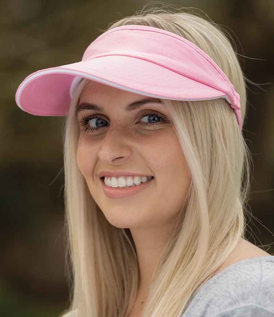 A person with long blonde hair is smiling and looking slightly left while wearing the Result Headwear Result Herringbone Sun Visor, which has a light pink color, pre-curved peak, and gray top. The blurred background is outdoors.