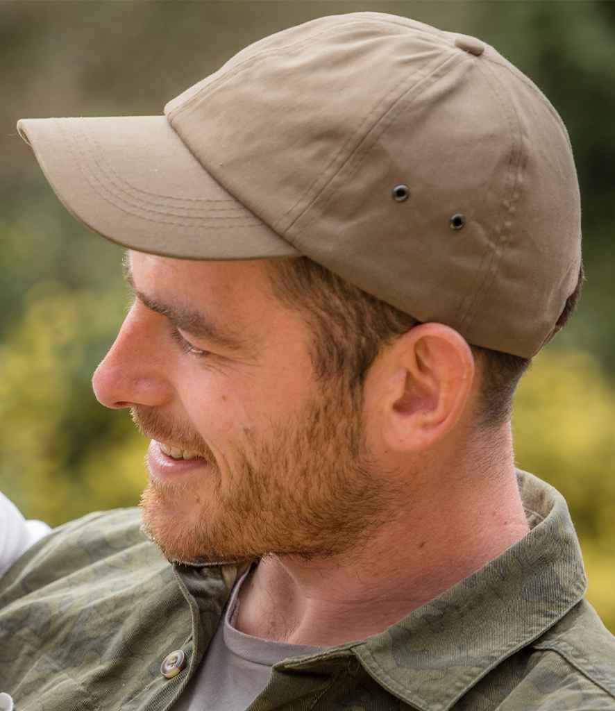 A man with a trimmed beard and short hair is smiling to the left, wearing a khaki Result Plush Cap by Result Headwear and a green patterned shirt. The background features blurred greenery.