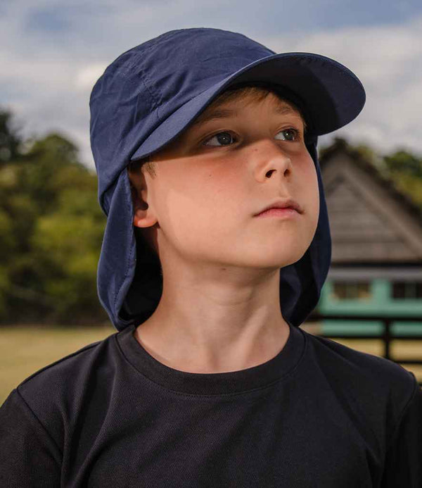 A boy in a black shirt and a Result Kids Legionnaires Cap by Result Headwear looks upward outdoors, with trees and a wooden structure blurred in the background.