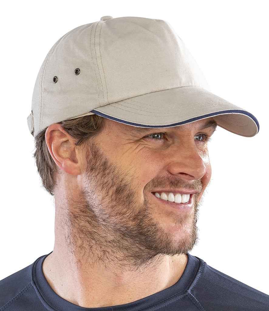 A man with light brown hair and a beard is smiling to the side, wearing a Result Headwear Result Plush Cotton 5 Panel Cap in light beige with an adjustable fabric buckle, along with a dark blue shirt against a plain white background.