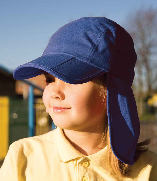 A child wearing the Result Kids Fold Up Legionnaires Cap by Result Headwear and a yellow polo stands outdoors in sunlight with eyes closed, looking slightly to the side; background is blurred.
