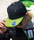 A person wearing the Result Headwear Result Contrast Bronx Snapback Cap in black and green, sunglasses, a black jacket with green lining, and a graphic tee looks down while adjusting the cap’s brim.