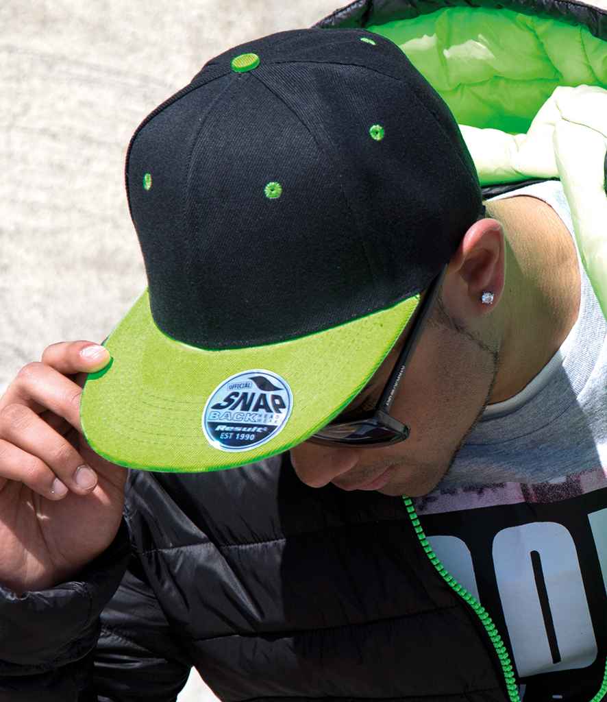 A person wearing the Result Headwear Result Contrast Bronx Snapback Cap in black and green, sunglasses, a black jacket with green lining, and a graphic tee looks down while adjusting the cap’s brim.