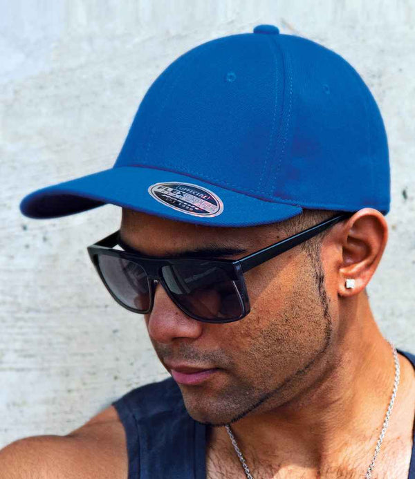 A person in a vibrant blue Result Kansas Flex Cap by Result Headwear, with stitched eyelets, black sunglasses, a black tank top, and earrings, faces slightly downward against a light-colored textured wall.