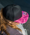 A person with long, light brown hair wears the Result Bronx Glitter Snapback Cap by Result Headwear. The pink marbled brim and circular patch stand out from above, and the stylish flat peak hat complements their dark outfit perfectly.