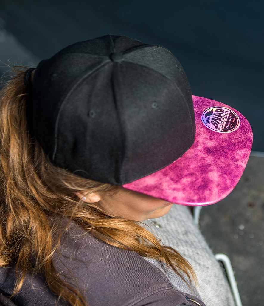 A person with long, light brown hair wears the Result Bronx Glitter Snapback Cap by Result Headwear. The pink marbled brim and circular patch stand out from above, and the stylish flat peak hat complements their dark outfit perfectly.