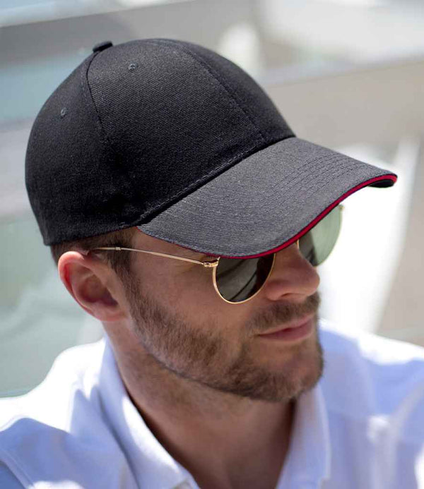 A person wears a Result Headwear Result Memphis Sandwich Peak Cap in black with a contrast peak, gold-rimmed sunglasses, light facial hair, and a white collared shirt.