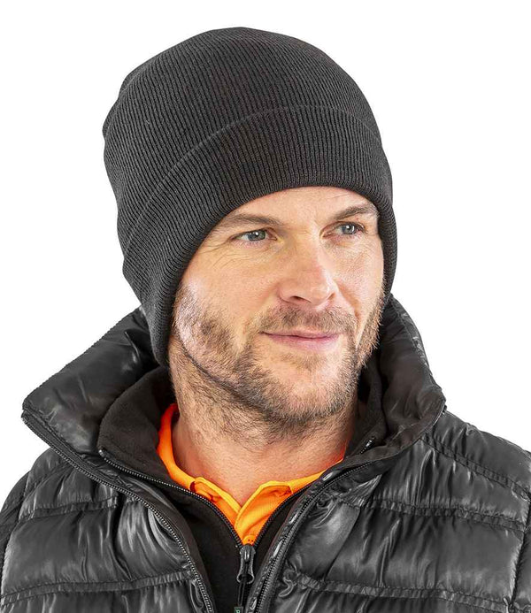 A man with light facial hair wears a Result Recycled Woolly Ski Hat, a shiny black puffer jacket, and an orange collared shirt. He looks slightly to the right with a neutral expression.