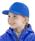 A young girl wearing the Result Recycled Junior Printers Cap by Result Recycled with a pre-curved peak and a blue puffer jacket smiles and looks to the side against a plain white background.